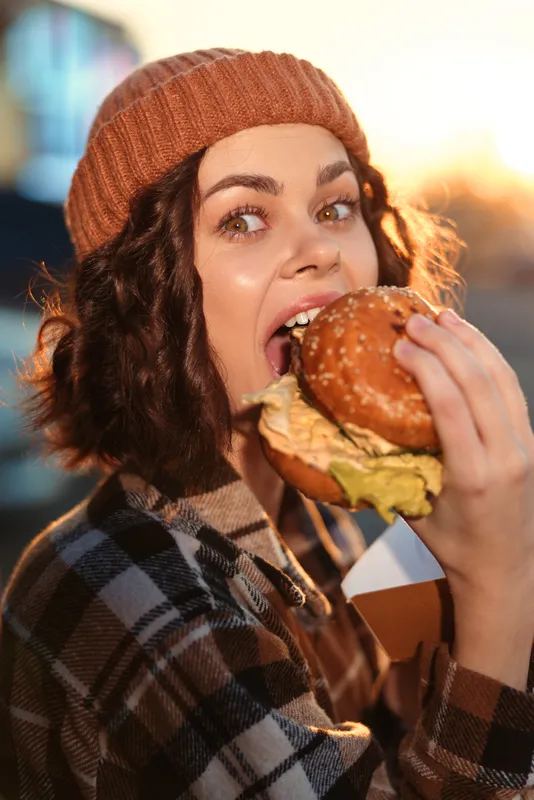 Woman eating burger in candid outdoor portrait during golden hour, cozy beanie and plaid jacket, authentic lifestyle moment with emotional storytelling and mindful living vibe.