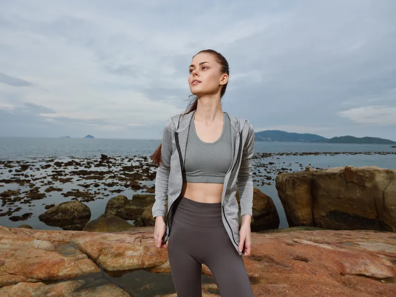 Young athletic woman in activewear on rocky beach, showcasing confidence and determination against a cloudy sky Fitness and outdoor lifestyle