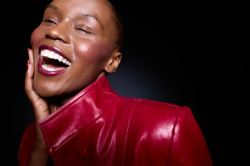 Close-up of a joyful dark skinned African woman laughing with eyes closed wearing a red leather jacket. Black background highlights her vibrant mood and radiant smile. Concept of happiness.
