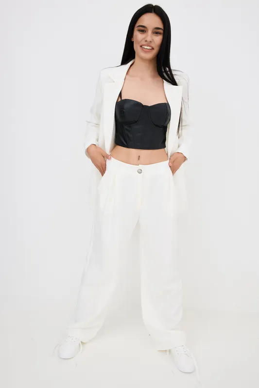 smiling woman wearing white wide pants and blazer with black crop top stands confidently with hands in pockets against a white background, modern fashion style, casual look