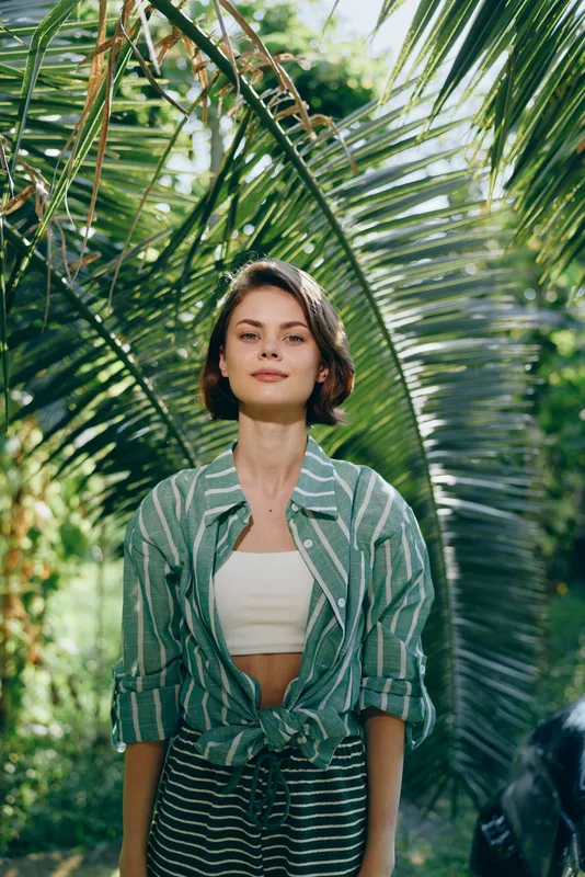 Portrait of a young woman standing amid lush tropical palm leaves, wearing a striped green jacket and white crop top.