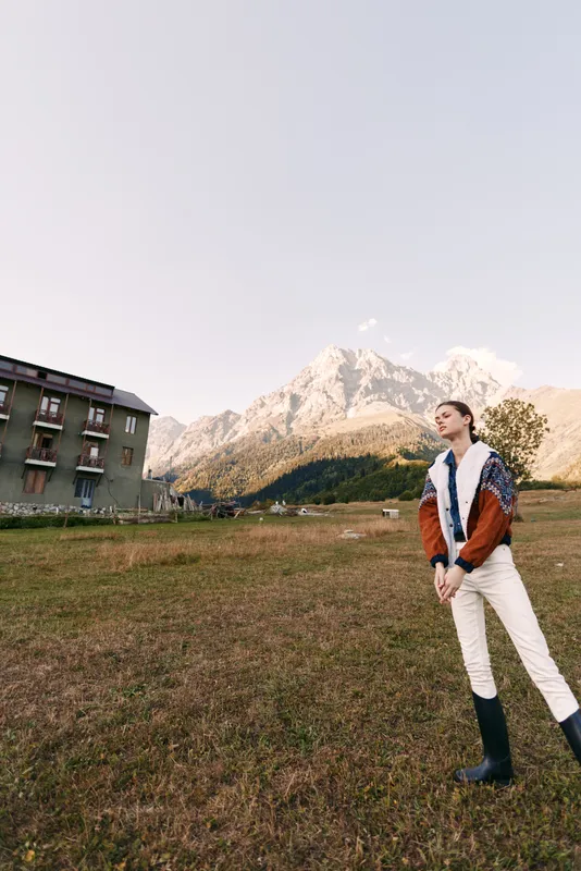 Woman in a jacket and boots standing in a meadow near a rustic building with alpine mountain backdrop, outdoor portrait capturing scenic landscape, sky and peaceful countryside view.