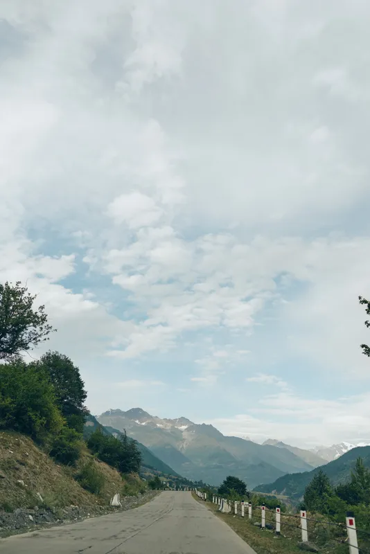Scenic mountain road winds through a rugged landscape under a bright sky, with trees along the roadside and distant peaks creating a peaceful travel scene.