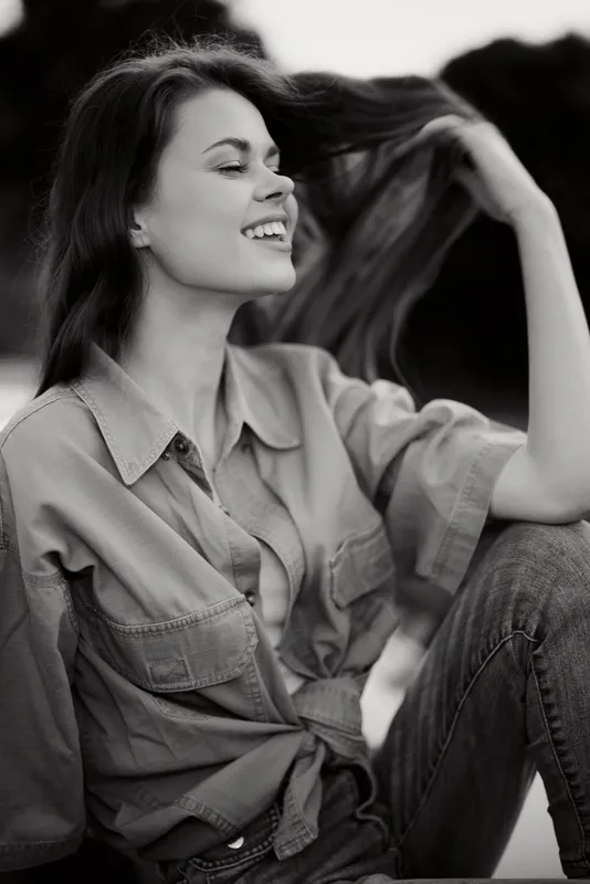 A carefree young woman enjoying the moment while playfully touching her hair, dressed in a relaxed outfit, set against a blurred outdoor background.