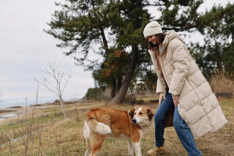 A woman in a puffy winter coat and beanie interacts with a dog in an outdoor park setting, capturing a cozy winter moment among grass, trees and a distant lake on a calm day
