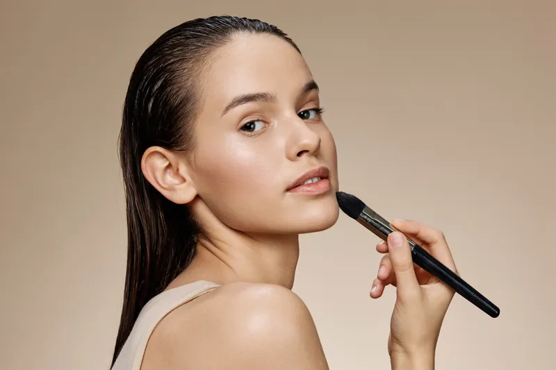 Young woman with natural makeup applying foundation in a beauty salon setting, showcasing healthy skin and confidence