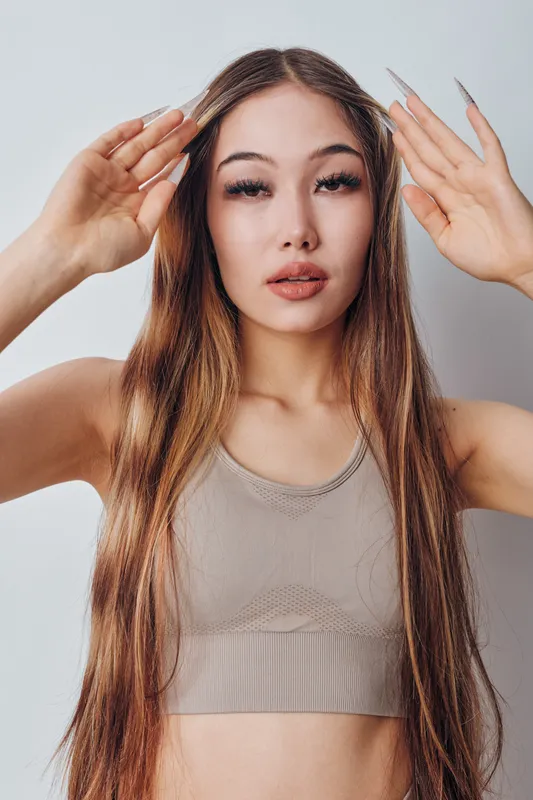 Woman with long straight hair and neutral makeup posing with hands near face wearing sleeveless top in studio. Portrait of young female model with calm expression and natural look.