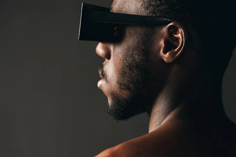 Creative conceptual portrait of African man wearing futuristic black visor glasses in visual style with dark background and dramatic lighting for modern technology theme.