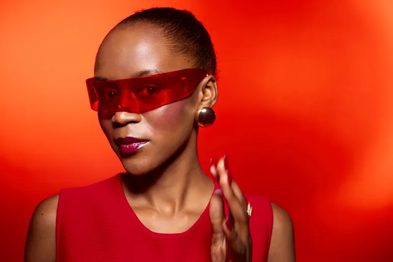 Confident African woman with dark skin wearing red sunglasses and red dress poses against vibrant red background. Modern fashion concept with bold accessories and stylish look.
