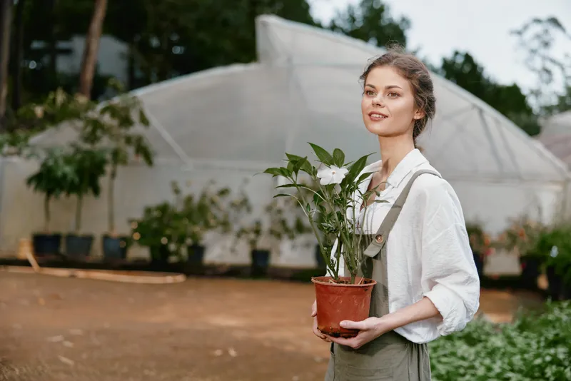 Young woman in casual attire holding a potted plant in a greenhouse setting, smiling amidst greenery, showcasing healthy gardening and nature connection