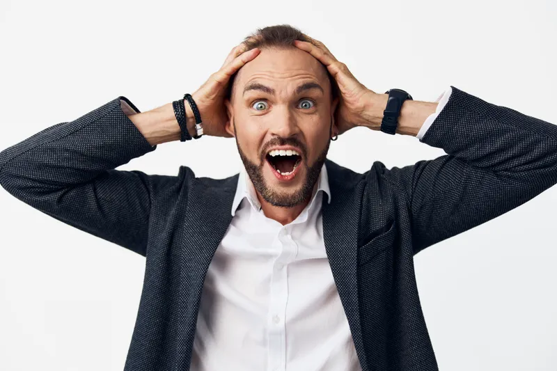 Professional man in business attire stands against a bold colored backdrop, showing surprise with hands on his head, conveying strong emotion and confident, modern presence.