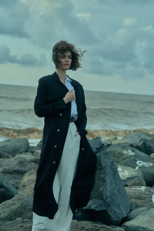 Woman in a long black coat on rocky shore, wind-swept hair, wearing business attire, by a moody sea backdrop, fashion and resilience.
