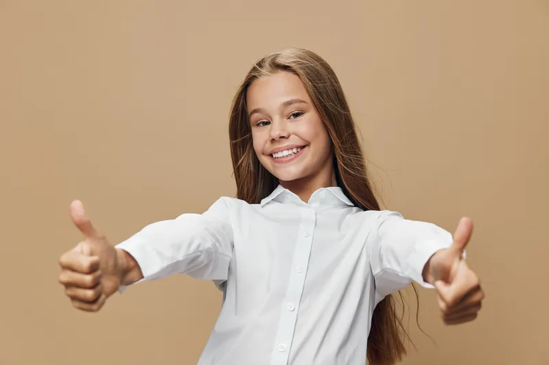 Smiling girl in white shirt showing thumbs up gesture with both hands, positive expression and casual look on beige background. Happy child with long hair and confident pose.