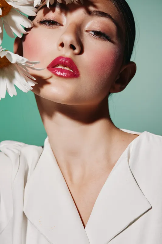 Young woman with bright pink glossy lipstick poses creatively with white flowers near face wearing white expert fashion outfit on green background in vibrant beauty concept.