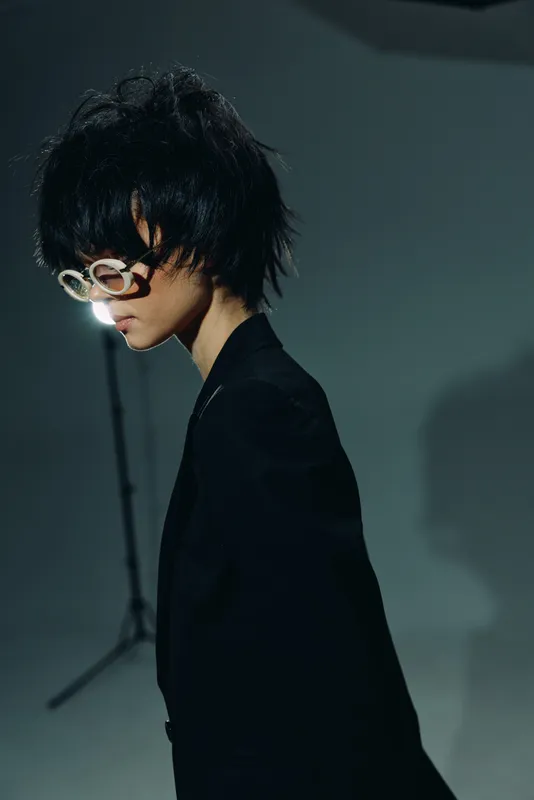 Young man with modern glasses in black outfit posing in moody studio lighting, dark background, stylish and creative portrait.
