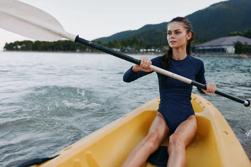 A female kayaker paddles a bright yellow kayak across calm water, wearing a dark wetsuit, focused expression, with gentle breeze and distant mountains in the background.
