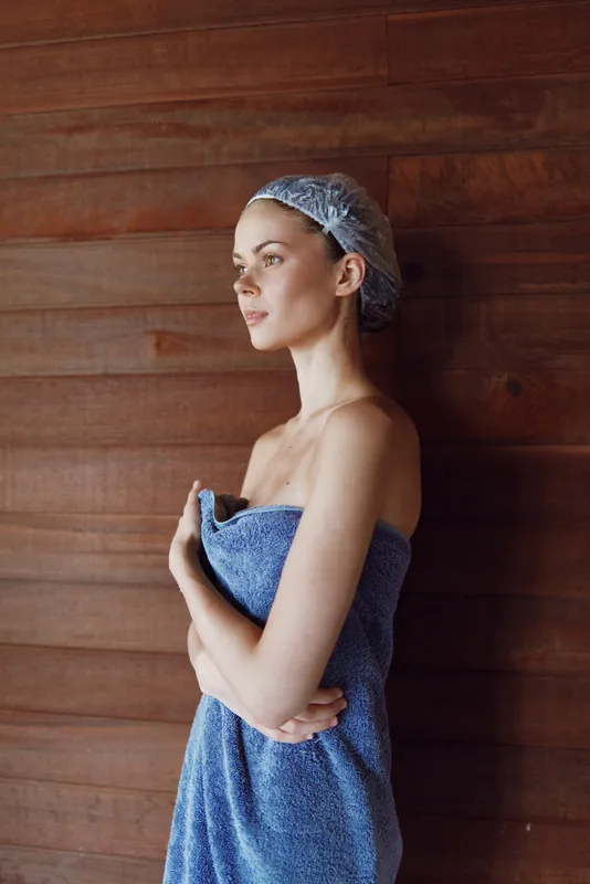 young woman wrapped in blue towel with shower cap standing contemplative against wooden wall in spa environment, calm and relaxed mood, natural light