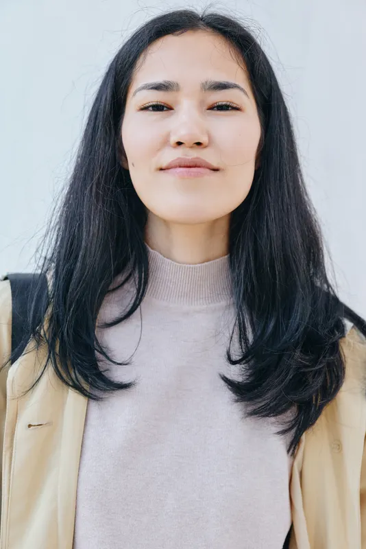 Female with shoulder length dark hair wearing a light sweater and beige jacket stands against a light wall, calm confident mood, minimal backdrop, natural lighting, stylish everyday vibe for