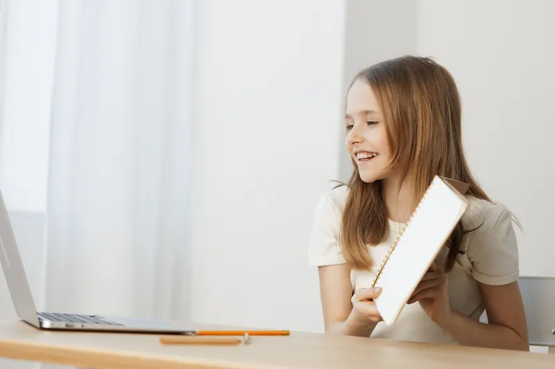 Young girl with long hair smiling while holding a book and using a laptop, bright and modern workspace, conveys joy of learning and exploration