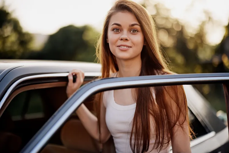 Young woman with long hair in a white tank top, smiling and leaning out of a car against a sunlit natural background, conveying joy and freedom during summer.
