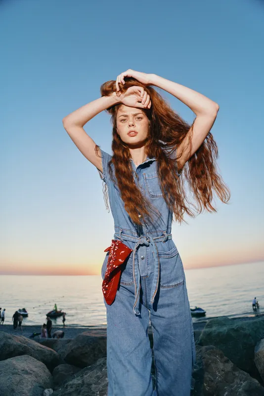 Modern boho-western denim street style woman posing confidently with red bandana by the sea at sunset in film color lifestyle setting.