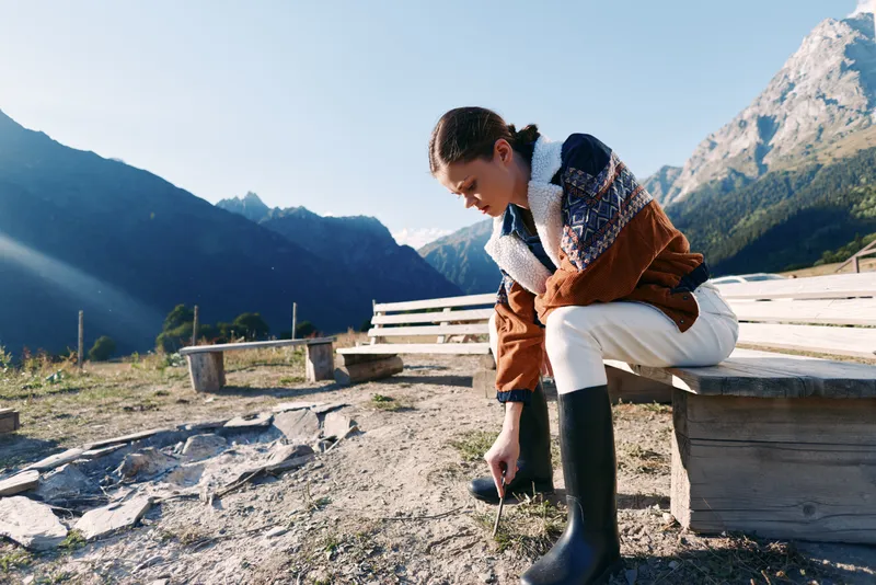 Woman in mountains sits on wooden bench wearing shearling jacket and rubber boots outdoors, adjusting footwear on a sunny alpine trail by rocky ground with panoramic valley view.