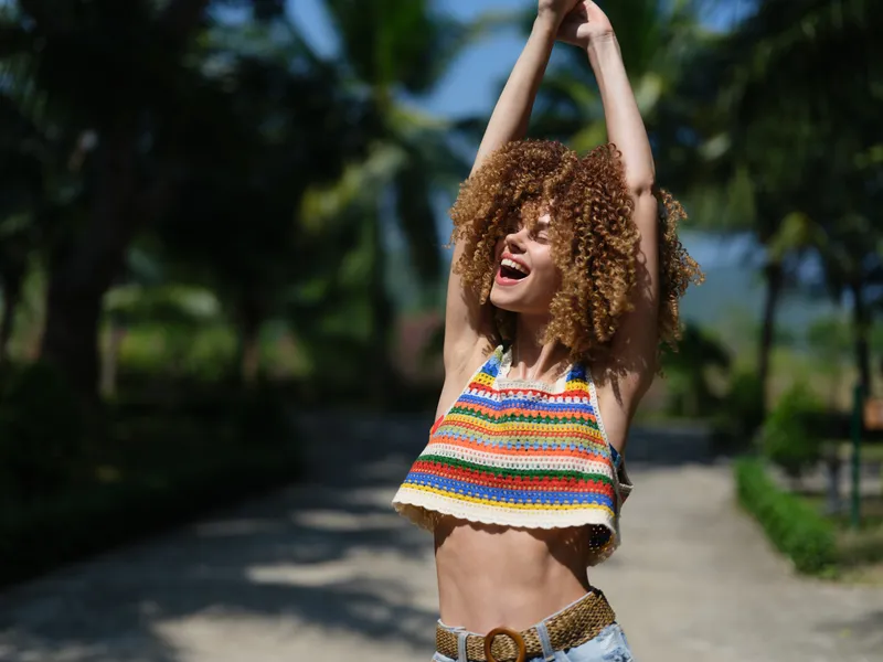 joyful young woman with curly hair wearing colorful striped crop top stretching arms outdoors, enjoying warm sunny day, tropical background with palm trees