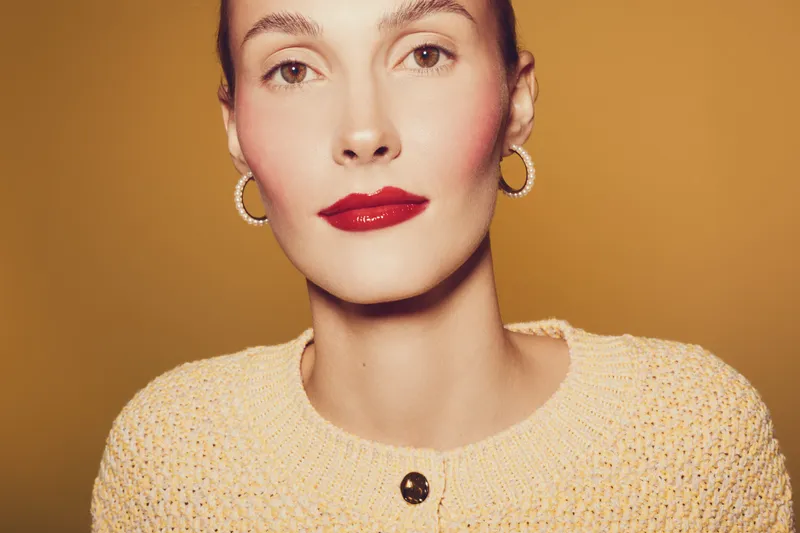 Close-up portrait of a woman with red lipstick and hoop earrings wearing a knitted sweater on a warm brown background. Concept of beauty, fashion, and elegant makeup style.