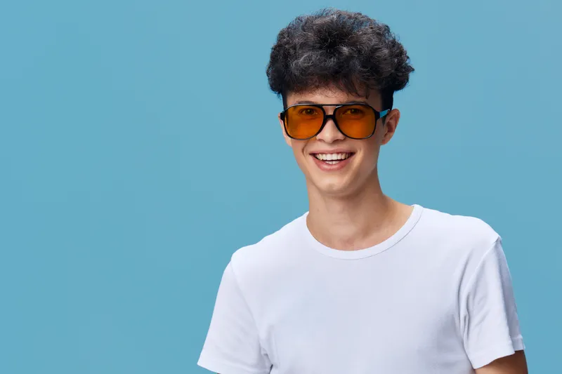 smiling young man wearing a white t-shirt and orange sunglasses, curly hair and cheerful expression, isolated on blue background, casual style