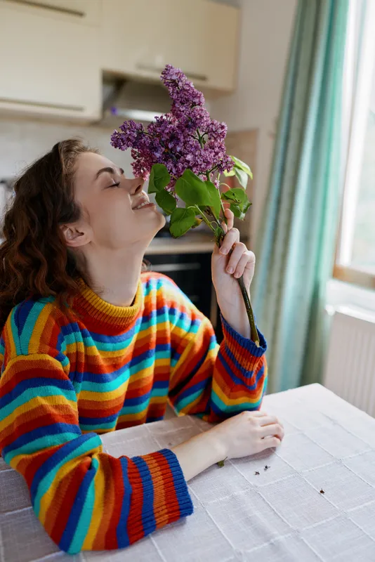 Cheerful woman indoors, sniffing purple flowers on a table in a bright kitchen, colorful striped sweater, cozy home ambiance, candid lifestyle moment
