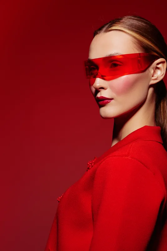Stylish woman in red jacket and red transparent glasses poses on red background. Fashion portrait with bold makeup and sleek hairstyle. Concept of modern style and confidence.