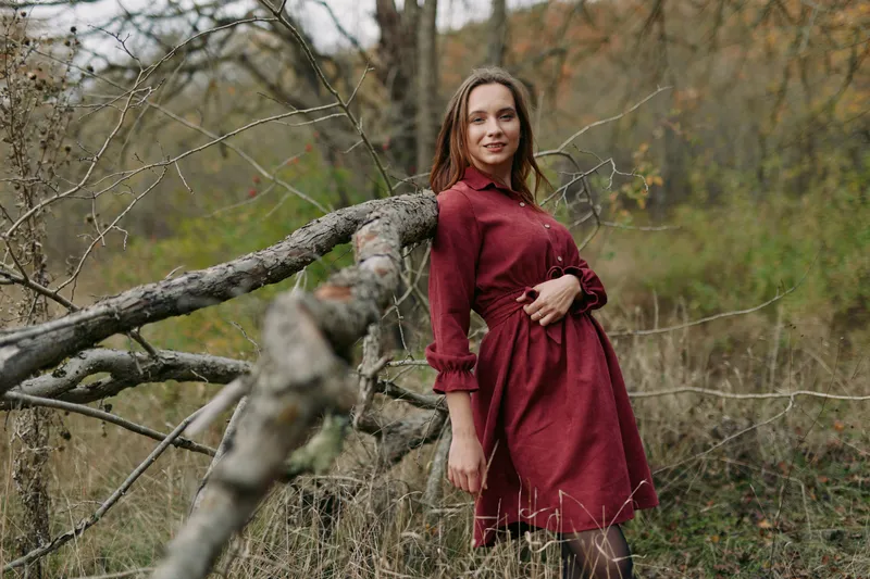 A woman stands in a muted autumn field wearing a burgundy dress, leaning against a fallen branch to convey authentic charm, quiet grace, and natural beauty in soft light.