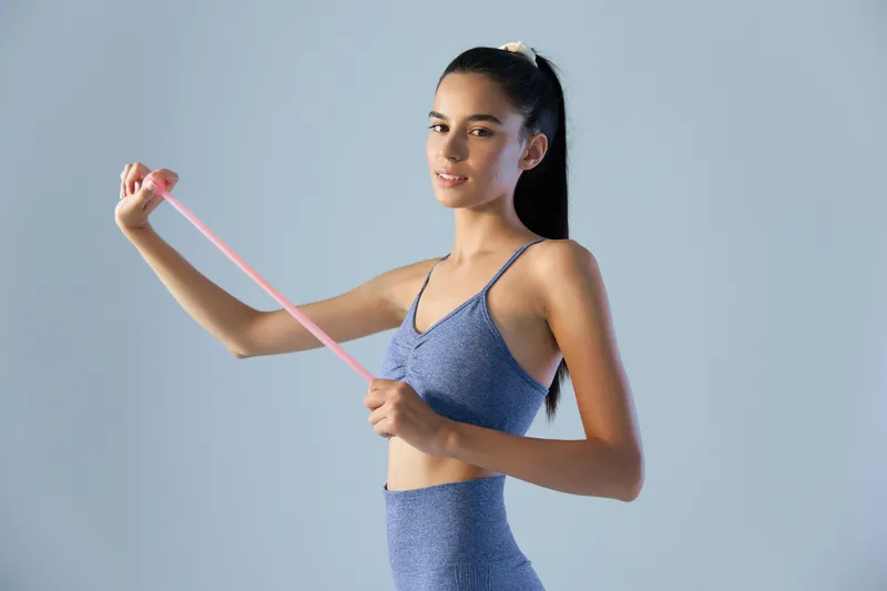 Woman sport fitness stretching with resistance band in blue activewear, demonstrating strength and flexibility against plain light blue background.
