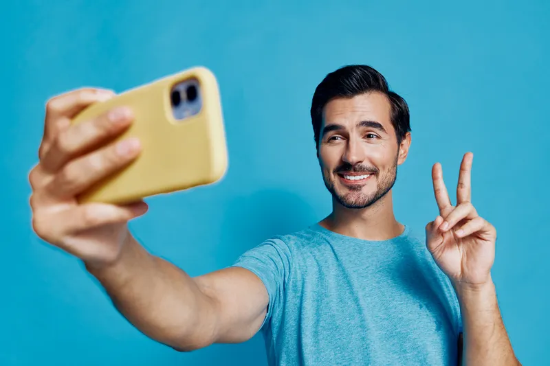 Smiling man taking a selfie with smartphone, showing peace sign, wearing casual blue t-shirt, standing against a vibrant turquoise background. Fun lifestyle concept.