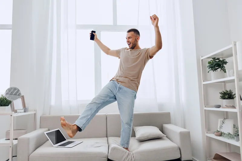 Happy man celebrating a digital detox moment while balancing on a couch, surrounded by sunlight, showcasing the blend of work and personal life.
