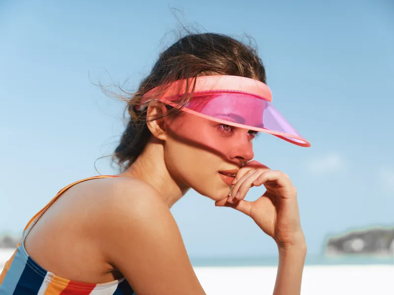 A young woman wears a pink visor and gazes toward the horizon on a sunny beach, creating a vivid portrait with warm light, smooth skin, and relaxed posture.