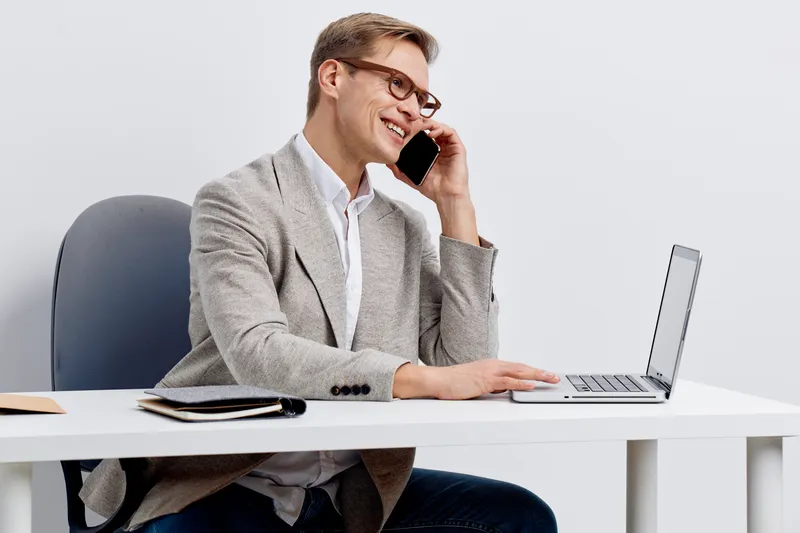 smiling man talking smartphone sitting at desk using laptop casual wear glasses modern workspace digital nomad concept