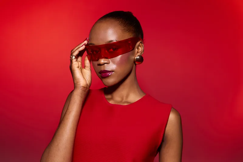Elegant African woman with dark skin wears red sleeveless dress and futuristic red visor glasses, posing confidently against a vibrant red background. Concept of fashion and style.
