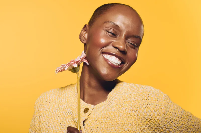 Happy African woman with dark skin smiles with closed eyes holding pink flower on yellow background. Concept of joy, beauty, natural skincare, and positive emotions in studio portrait.