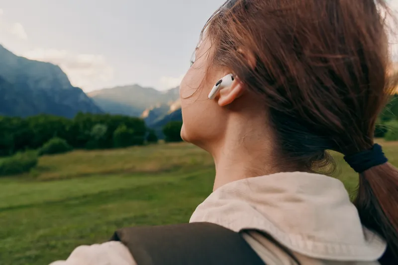 A woman with headphones stands in open countryside, overlooking distant mountains, capturing a relaxed outdoor lifestyle moment in fresh air and wide landscapes.
