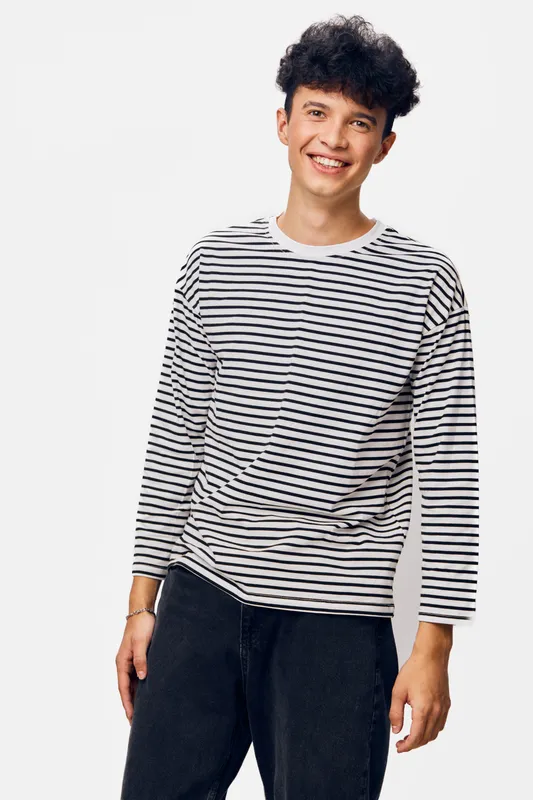 smiling man wearing a striped long sleeve shirt and black pants, standing against a plain white background, casual style with curly hair, happy expression