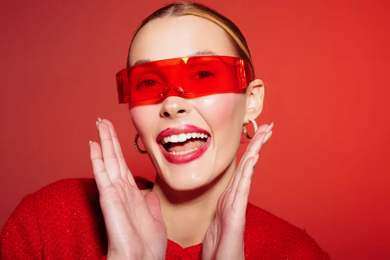 Happy young woman with red lipstick and red sunglasses smiles widely on red background. Fashion portrait with bright makeup and joyful expression. Concept of style and confidence.