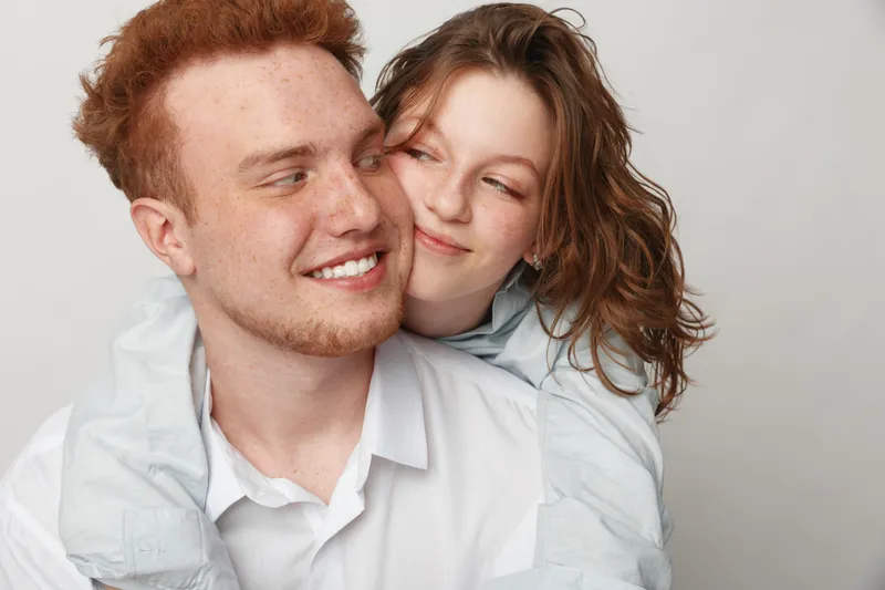 Happy young couple with red hair embraces and smiles in casual white clothing. Close affectionate moment with natural expressions on neutral background. Concept of love and friendship.