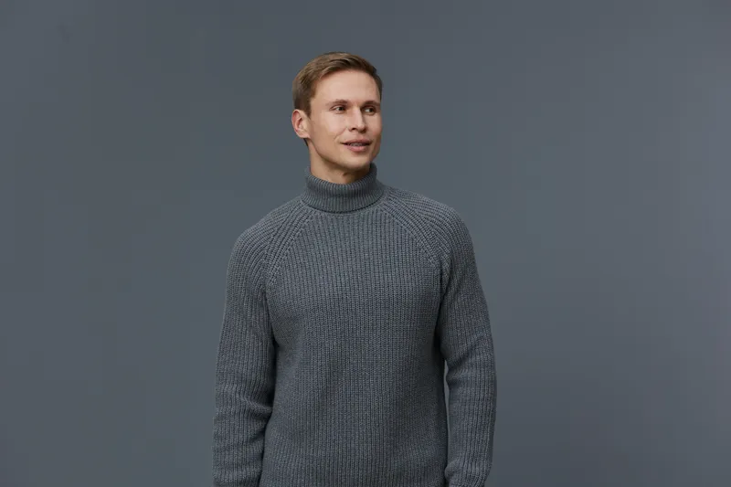 Man wearing gray knitted turtleneck sweater posing confidently against plain gray background. Casual fashion, winter clothing, male model, fashionable style, portrait, indoor studio shot.