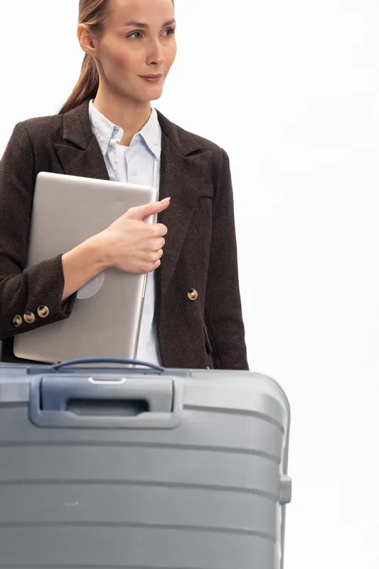 Businesswoman in blazer holds laptop and stands behind suitcase on white background. Concept for travel, business trip, work, and professional lifestyle in modern corporate environment.