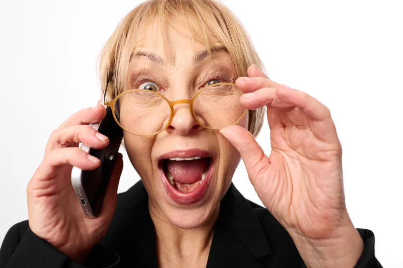 Senior woman with glasses on phone, surprised call, joyful communication, business suit, studio portrait, isolated on white, hands holding cellphone, open mouth expression