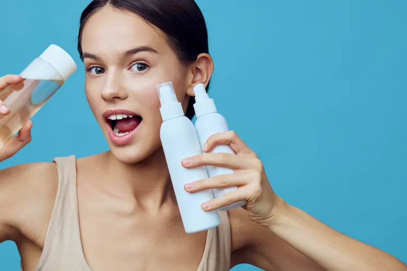 Young woman holding skincare products, excited expression, light background, presenting beauty routine for healthy skin