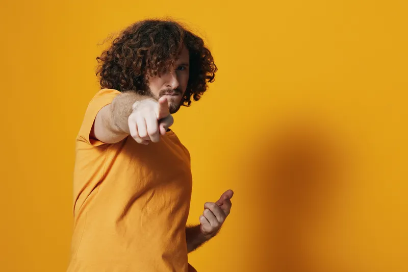 Young man with curly hair in orange t shirt on bright yellow background, expressing confidence and positivity while pointing forward, summer vibes