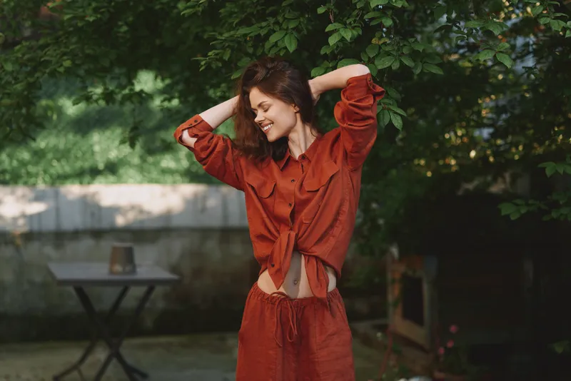 A woman in an orange casual outfit stretches her arms and smiles outdoors, sunlight in a garden setting, conveying relaxed, carefree lifestyle