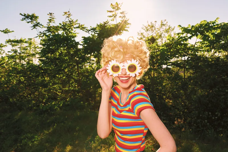 A young woman in a striped shirt playfully wears oversized flower sunglasses in a sunlit outdoor setting, radiating a retro, carefree mood.